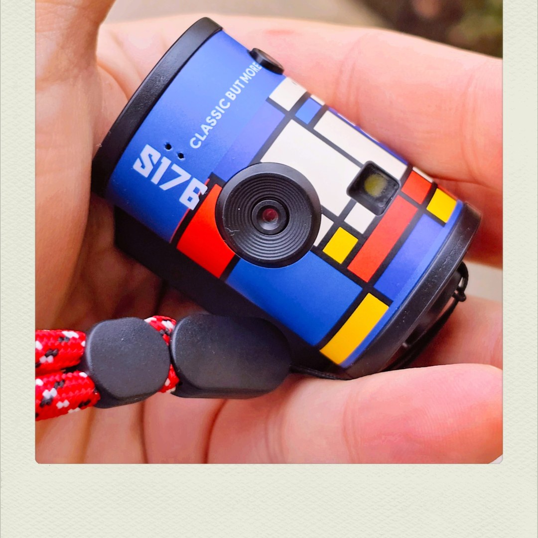 A close-up of a hand holding a small, cylindrical toy camera decorated with a Mondrian-style geometric pattern of blue, red, yellow, and white sections. The text 'S176' and 'CLASSIC BUT MORE' is printed on the blue section near the lens. A red-and-white patterned wrist strap is attached to the camera.