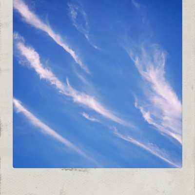 Wispy white clouds stretch diagonally across a vibrant blue sky, framed by a weathered white Polaroid border.