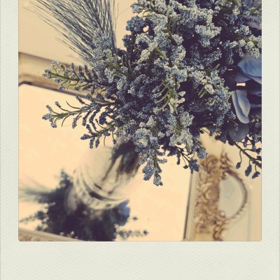 Top-down view of blue sprigs in a glass vase on a mirror, reflecting the bouquet and an ornate frame.
