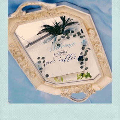 Cream-colored ornate tray mirror reflecting a white wedding sign with the text "Welcome to our happily ever after" in blue script, set against a light blue background.