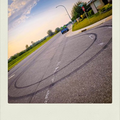Dutch-angle shot of circular black skid marks on a grey asphalt street corner, with a blue truck parked in the distance under a pale blue sky.