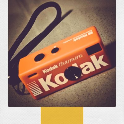 A close-up of a small, bright orange Kodak Charmera keychain digital camera with a black wrist strap, set against a grey background with a yellow stripe inside a Polaroid frame.