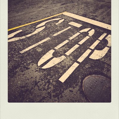 High-angle view of stenciled "STOP" road marking on textured dark asphalt with a yellow line and manhole cover, styled with a warm vintage Polaroid border.