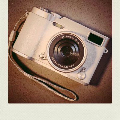 Top-down view of a silver compact digital camera with a wrist strap on a brown textured background. Lens text reads "16X Powerful Zoom" and "F/2.2 f=3.37mm Auto Focus".