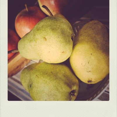 A close-up photograph of three speckled green and yellow pears in the foreground, with red apples nestled behind them in a soft-focus background.