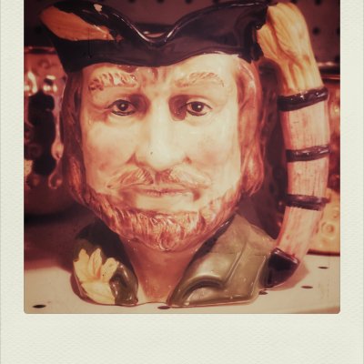 A close-up photograph of a ceramic Toby jug shaped like a bearded man's head, wearing a dark cap and a green collar, displayed on a shelf.