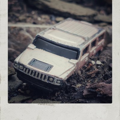 A close-up, high-angle shot of a miniature toy Hummer SUV, beige and weathered, wedged into dark soil and wood chips. The toy car is angled towards the left, half-buried in the rough terrain, captured inside a textured, vintage Polaroid-style frame.