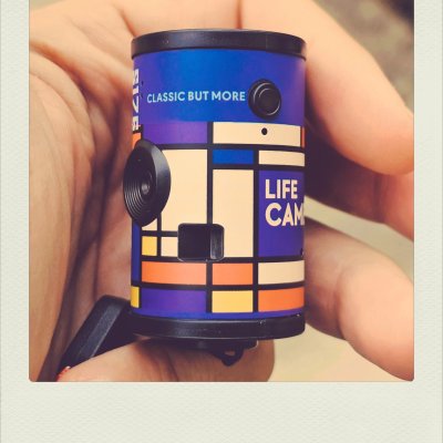 A hand holding a small cylindrical camera decorated with a colorful geometric pattern of blue, yellow, orange, beige, and purple blocks separated by black lines. Text on the camera body reads "CLASSIC BUT MORE" and "LIFE CAM" in white. The photo is enclosed in a classic white polaroid-style frame.
