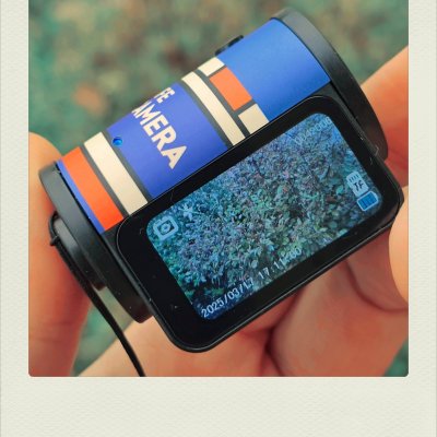 A close-up of a hand holding a small cylindrical digital camera with a retro blue, orange, and cream-striped design. The camera body features the word 'CAMERA' in white, while the rectangular LCD screen on the back shows a live preview of green leafy bushes, complete with interface icons and the date '2025/03/17'.