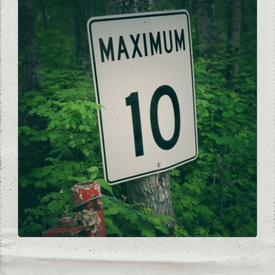 A white road sign with black text reading "MAXIMUM 10" is mounted on a tree trunk in a dense green forest. The sign is surrounded by lush green foliage, with a rusted red metal post visible in the lower-left corner. The entire image is presented in a white polaroid-style frame.