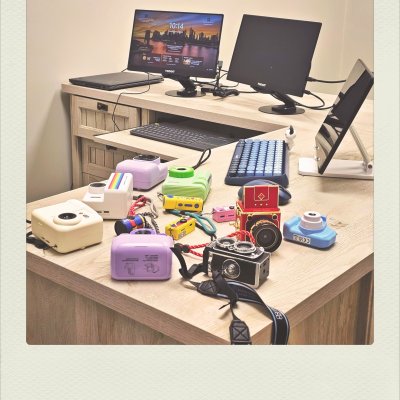 A high-angle shot of a light-wood L-shaped desk holding various colorful toy and instant cameras in pastel purple, green, yellow, red, and blue. In the background, a dual-monitor computer setup, laptop, and tablet are neatly arranged on the office desk.