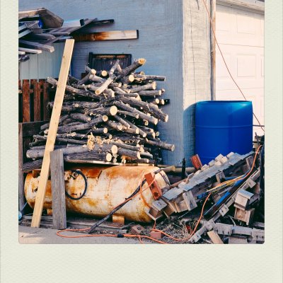 A stack of cut tree branches piled atop a rusty, weathered yellow cylinder tank. Next to them stands a vibrant blue plastic barrel against a light-grey concrete wall, with scrap wood and orange cables scattered below.