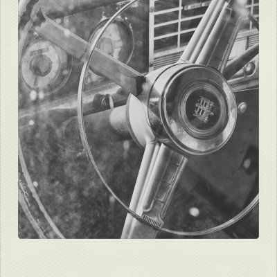 A black and white close-up shot of a vintage car steering wheel with a circular metal horn ring and central emblem, viewed through a dusty, speckled window pane. In the blurred background, the retro dashboard and speedometer are partially visible.