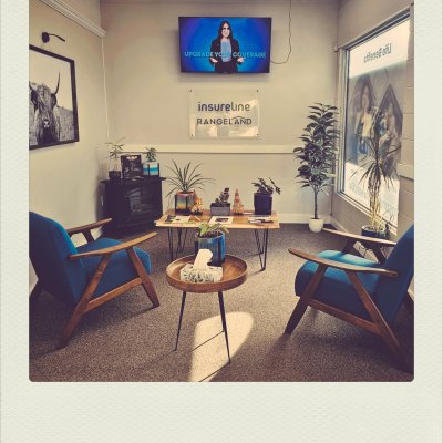 A warm office reception room with two blue mid-century modern armchairs facing a wooden coffee table decorated with plants. On the wall, a sign reads "insureline RANGELAND" below a TV displaying an ad with a woman and the words "UPGRADE YOUR COVERAGE". A framed black-and-white print of a highland cow hangs on the left wall.