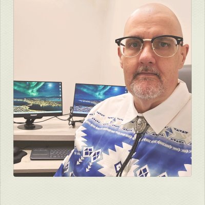 A portrait faux-Polaroid of a bald man with a goatee and black-rimmed glasses, looking at the camera. He is wearing a white collared shirt with a blue geometric print and a silver-toned bolo tie. Behind him, two computer monitors display an aurora borealis landscape on a desk.