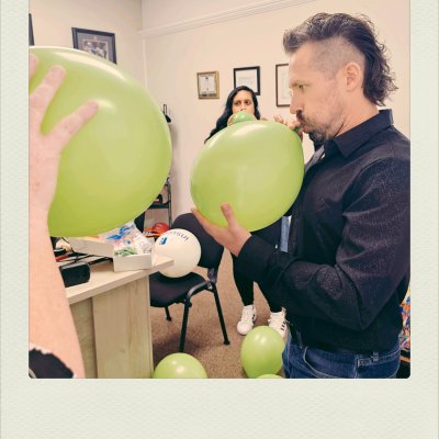 A man with a mullet haircut and black shirt blows up a lime green balloon in an office room. A hand in the foreground holds a large green balloon, and other green and white balloons lie on the floor and a chair.