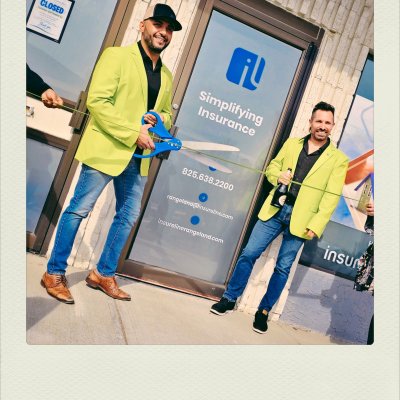 Two men in matching bright lime-green blazers and blue jeans stand outside an office door for a ribbon-cutting ceremony. The man on the left wears a cap and cuts a green ribbon with giant blue scissors, while the man on the right smiles and holds a bottle of champagne. The office door behind them displays a blue logo and the words "Simplifying Insurance."