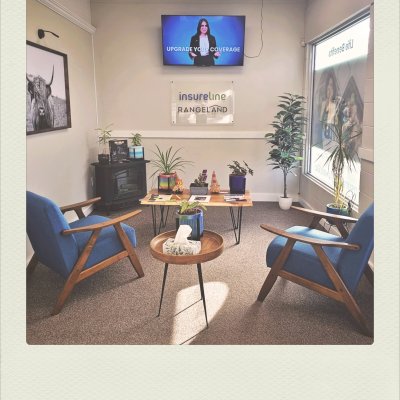 A well-lit office waiting room with two blue wooden-framed armchairs facing each other. Between them is a low coffee table with potted plants, and a small wooden side table with a tissue box sits in the foreground. On the back wall is a sign reading "insureline RANGELAND" below a TV displaying "UPGRADE YOUR COVERAGE" next to a presenter.