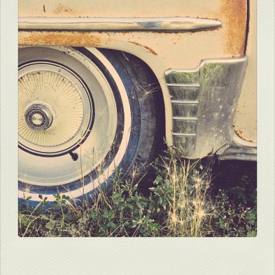 Close-up of a vintage cream-colored car's whitewall tire with a ribbed hubcap, partially obscured by overgrown green grass and dry weeds. The metal body above the wheel shows signs of rust along the chrome trim and fender.